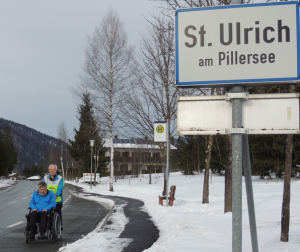 Deelnemer in rolstoel bij plaatsnaambord St.Ulrich am Pillersee